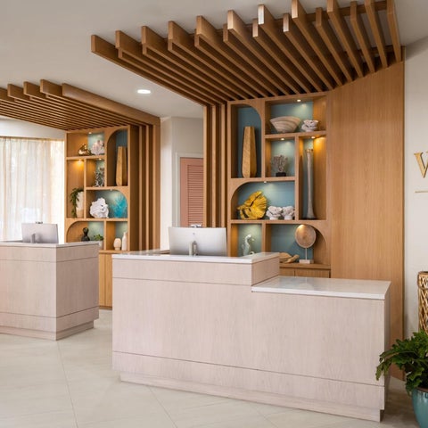 A modern hotel lobby features a reception desk, wooden shelving, and a large logo against a backdrop of natural light.