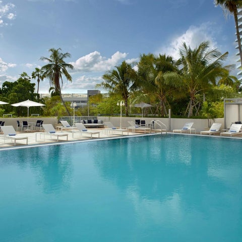 A large, rectangular swimming pool with numerous lounge chairs and umbrellas surrounds a lush, tropical landscape.