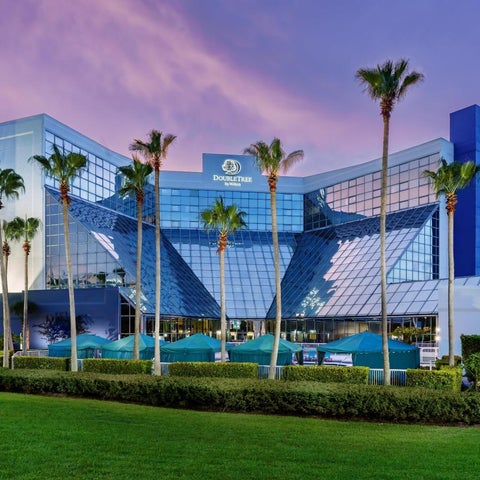 A large, modern hotel building with a glass facade, palm trees, and a manicured lawn in front.