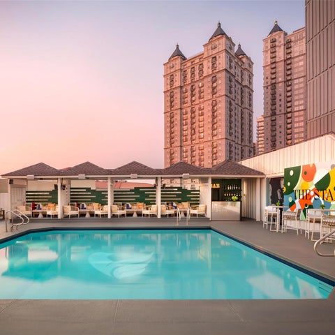A rectangular swimming pool with lounge chairs and a bar area sits in front of a large hotel building at dusk.