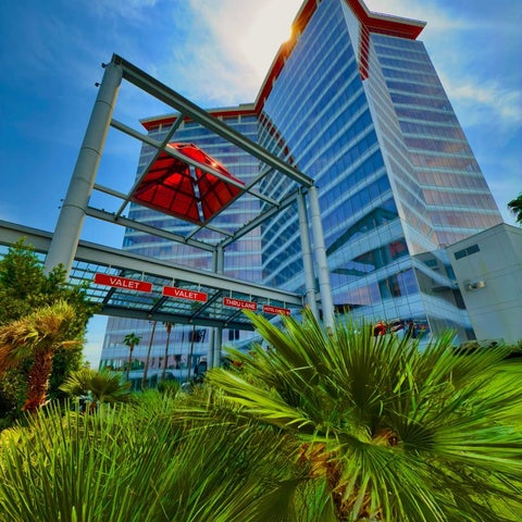 A tall, modern hotel building with a glass facade and a valet entrance is seen against a bright, sunny sky.