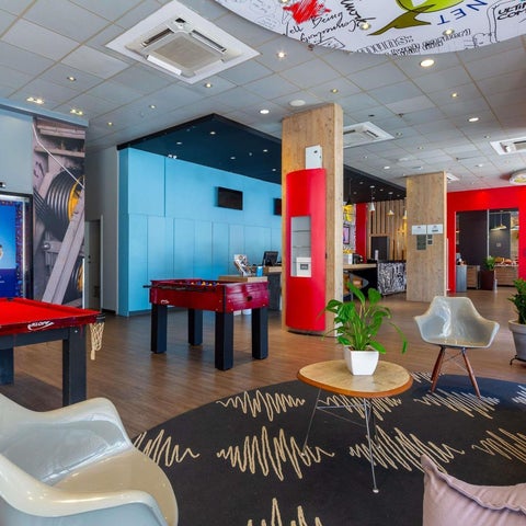 A modern hotel lobby features a pool table, lounge seating, and a reception desk with vibrant blue and red accents.