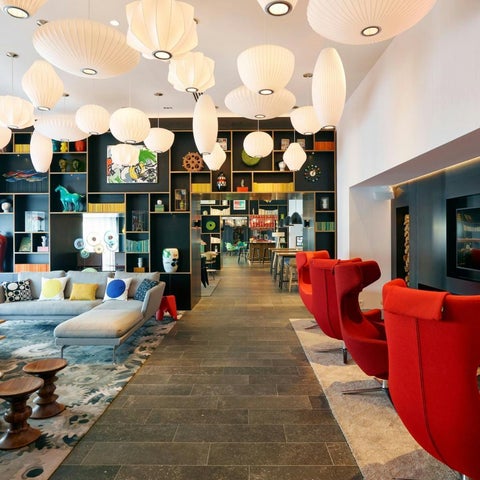 A modern lobby features suspended paper lanterns, a large sectional sofa, and vibrant red chairs with a dark blue accent wall.