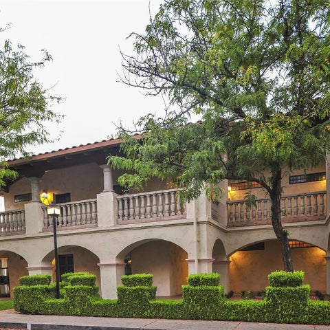 A multi-story building with a classical facade features arched walkways, a balcony, and manicured hedges in the foreground.