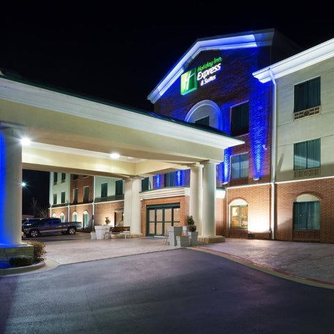 An exterior view of a brightly lit Holiday Inn Express & Suites building at night, featuring a covered entrance and brick facade.