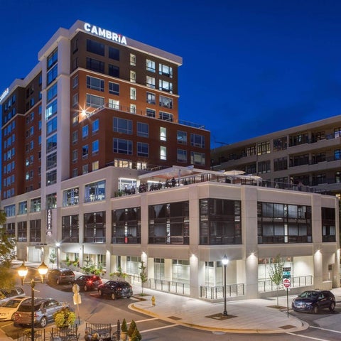 A modern hotel building with a brick and white facade is illuminated at dusk, featuring a rooftop terrace and street-level parking.
