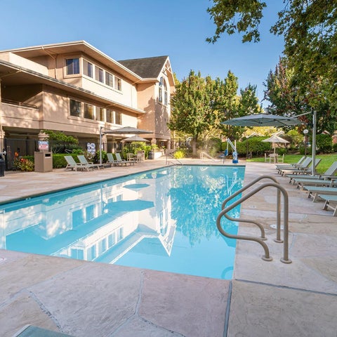 A rectangular swimming pool reflects a building with lounge chairs and umbrellas surrounding the pool area.