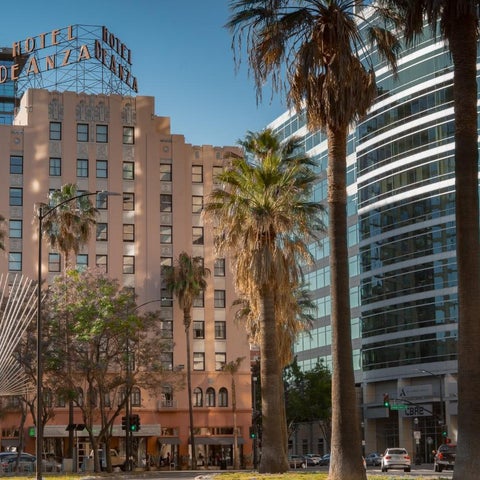 The Hotel de Anza building is shown with palm trees and other buildings in a sunny urban setting.