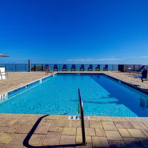 A bright blue pool with lounge chairs and umbrellas sits on a tiled patio overlooking a clear blue ocean.