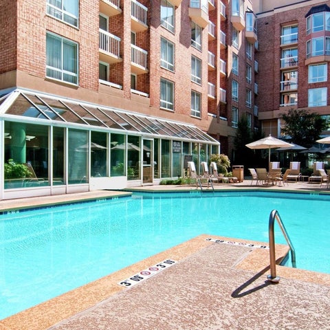 A rectangular swimming pool with a tiled deck and lounge chairs sits adjacent to a brick hotel building with a glass-enclosed patio.