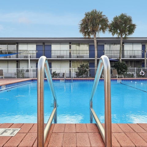 A bright blue pool with a tiled deck and metal ladder is situated in front of a hotel with palm trees.