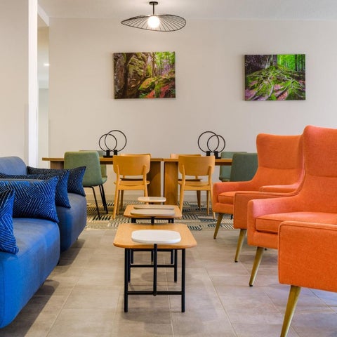 A modern lobby features a blue sofa, orange armchairs, and a wooden table with black chairs and artwork on the walls.