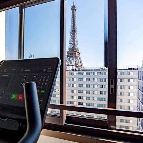 A treadmill sits in a room with a large window showcasing a view of the Eiffel Tower and surrounding Parisian buildings.