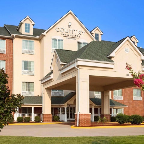 A Country Inn & Suites building features a covered entrance, brick facade, and landscaping with trees and shrubs.