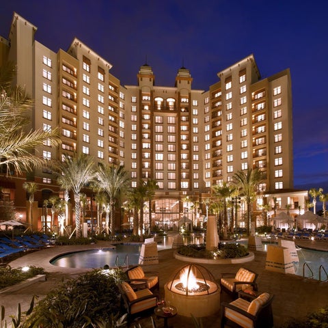A large, illuminated hotel building with a pool, lounge area, and palm trees in the foreground at dusk.