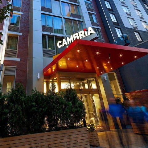 The Cambria hotel entrance features a red awning, glass doors, and lush greenery in front of a brick facade.