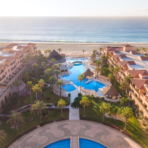 An aerial view showcases a sprawling resort with a large, turquoise pool, lush landscaping, and a sandy beach meeting the ocean.