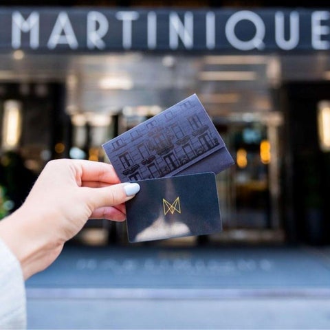 A hand holds two dark cards in front of the entrance of the Martinique hotel, with the building facade visible in the background.