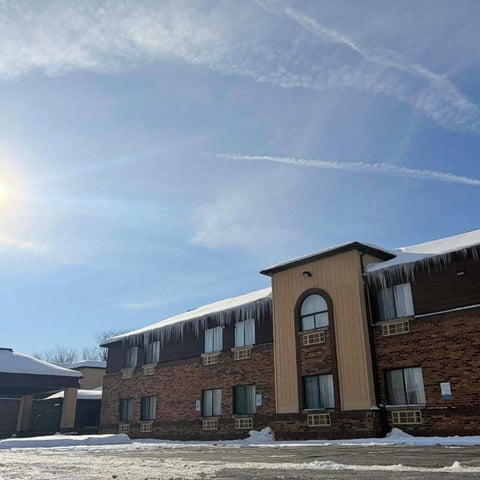 A brick hotel building with a flat roof and numerous windows stands against a bright, partly cloudy sky with contrails.
