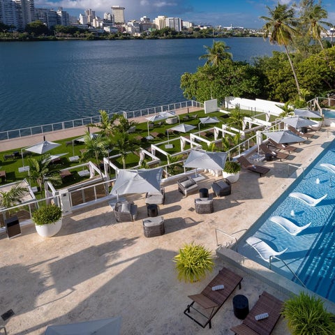 An aerial view showcases a resort pool area with lounge chairs, umbrellas, and a waterfront view of a city skyline.