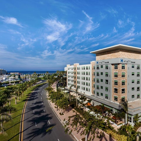 A Hyatt House hotel stands prominently alongside a tree-lined road and marina under a bright, partly cloudy sky.