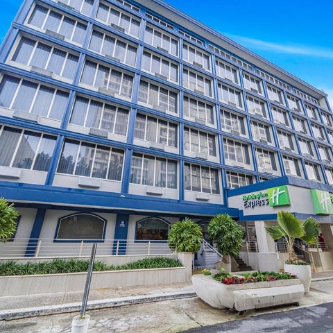 A multi-story hotel building with a blue facade and green landscaping is seen on a sunny day.