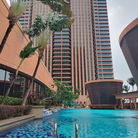 A vibrant blue pool with a tiled bottom sits in front of a tall, modern hotel building and lush tropical foliage.