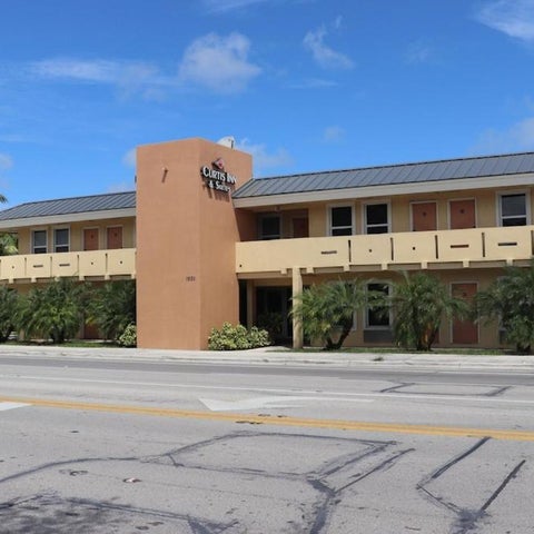 A two-story hotel building with a tan facade, a tall tower, and palm trees lining the front.