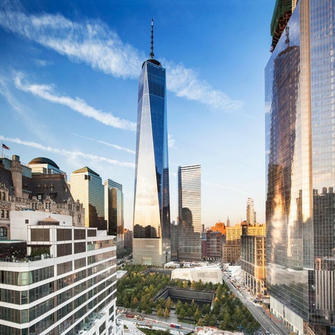 A wide view showcases One World Trade Center and surrounding buildings against a partly cloudy blue sky in New York City.