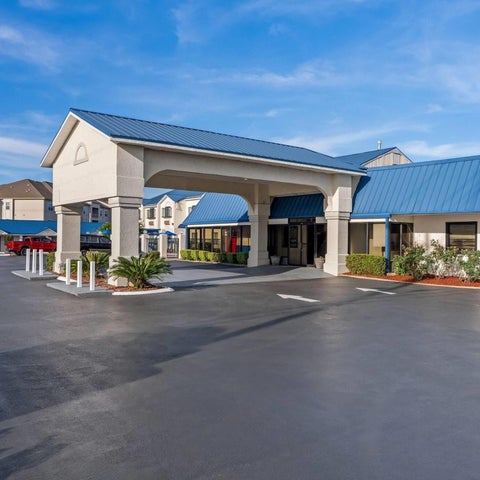 A wide shot shows the front of a hotel with a covered entrance, parking lot, and blue sky with scattered clouds.