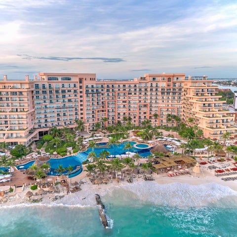 An aerial view showcases a large resort with multiple pools, a beachfront, and lush tropical landscaping.