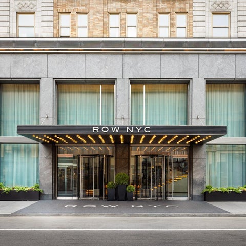 The entrance of Row NYC hotel features a marquee sign, glass doors, and a gray stone facade with planters.