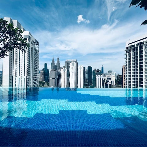 A vibrant blue infinity pool overlooks a cityscape of modern skyscrapers under a partly cloudy sky.