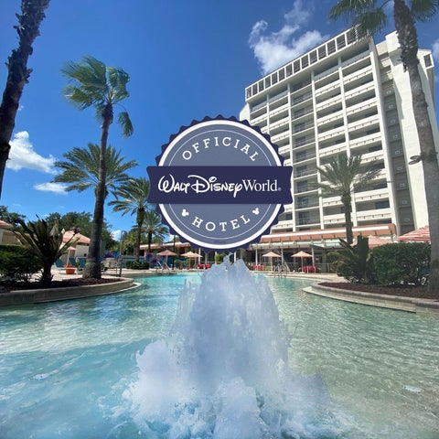 A large Disney hotel building is visible with a fountain and palm trees in the foreground, under a bright blue sky.