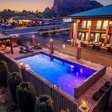 An elevated view shows a hotel pool with lounge chairs, a building facade, and a red rock formation in the background at dusk.