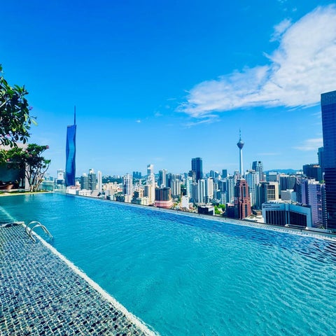 A vibrant blue infinity pool overlooks a cityscape featuring modern skyscrapers and the iconic Petronas Towers under a clear sky.