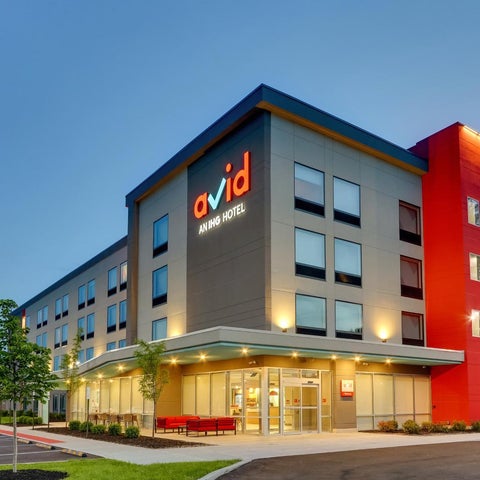 A modern, multi-story Avid Hotel building with a red accent tower and a parking lot in front of it.
