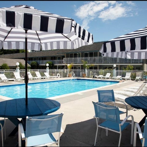 A poolside area features a blue pool, lounge chairs, tables with chairs, and striped umbrellas under a sunny sky.