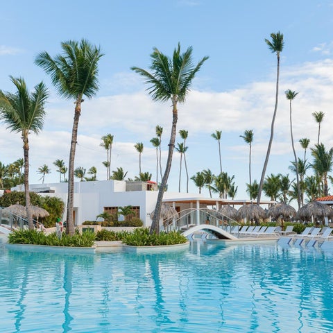 A large, turquoise pool with lounge chairs and palm trees surrounds a resort building with a thatched roof.