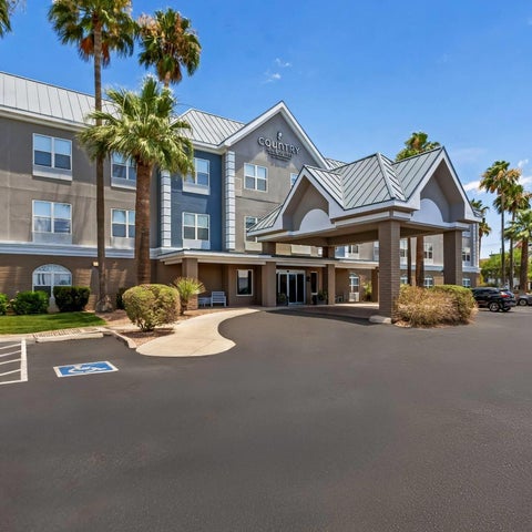 A multi-story hotel with a covered entrance and palm trees stands in front of a parking lot under a clear blue sky.