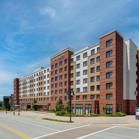 A large, multi-story hotel building with brick and light-colored facades sits near a road and a body of water.