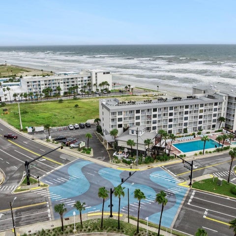 An aerial view shows a beachfront hotel complex with a pool, palm trees, and a busy coastal road in the foreground.