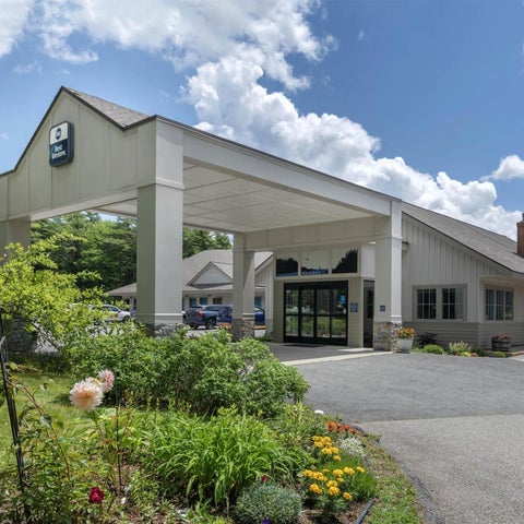 A modern hotel building features a covered entrance, landscaping, and a chimney against a partly cloudy sky.