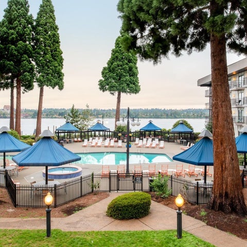 A hotel pool area features blue umbrellas, lounge chairs, a jacuzzi, and a view of a waterfront cityscape.
