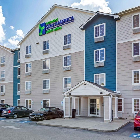 A StayAmerica hotel building features a light-colored facade with blue accents and a covered entrance with parking in front.