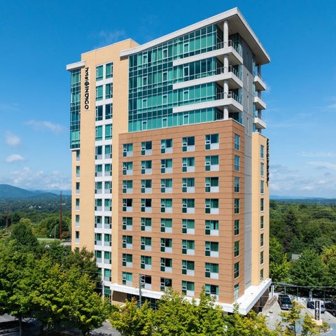 A tall, modern apartment building with a yellow and brown facade features balconies and overlooks a green landscape and distant mountains.