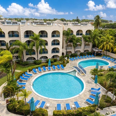 An aerial view showcases a resort with a curved swimming pool, lounge chairs, palm trees, and a multi-story building.