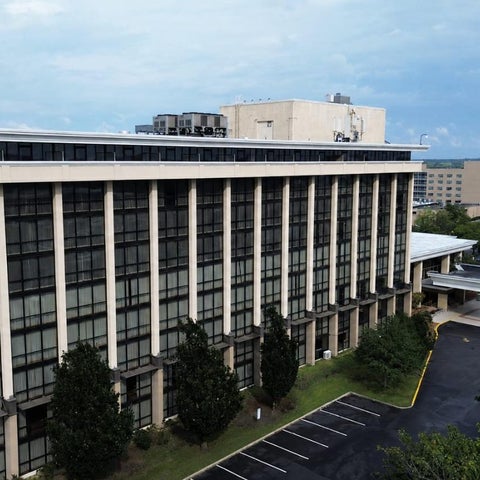 A large, multi-story building with a grid-like facade and surrounding greenery, featuring a driveway and parking area.