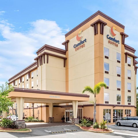 A multi-story Comfort Suites hotel building with a covered entrance and parking lot under a partly cloudy sky.