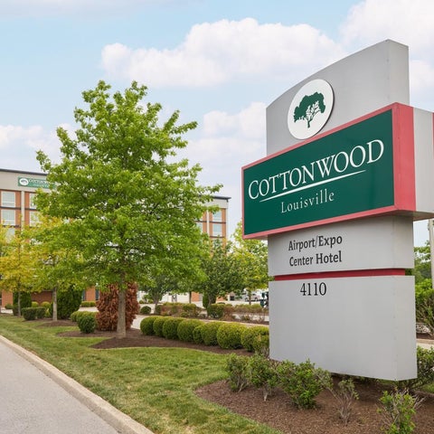A sign for the Cottonwood Airport/Expo Center Hotel stands in front of the building with lush greenery and a road.
